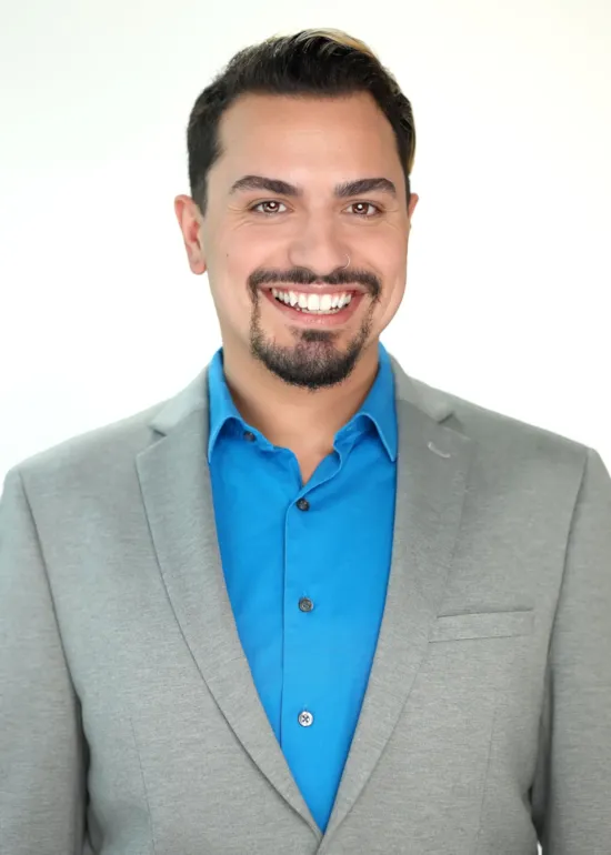 Smiling man with dark hair and goatee wearing a blue shirt and light gray blazer against a light background.