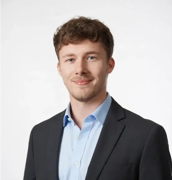 Man with blonde hair wearing a blue top with a dark suit, smiling in front of a plain white background.