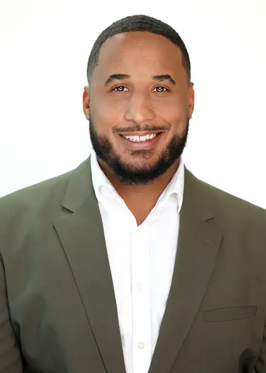 Smiling man with short hair and beard wearing a white shirt and olive green blazer against a plain background.