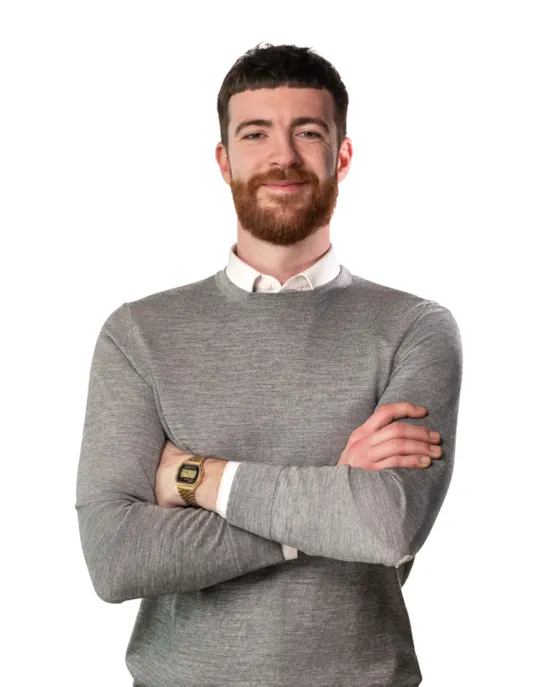 A smiling man with a beard stands confidently with arms crossed, wearing a grey sweater and a gold wristwatch.
