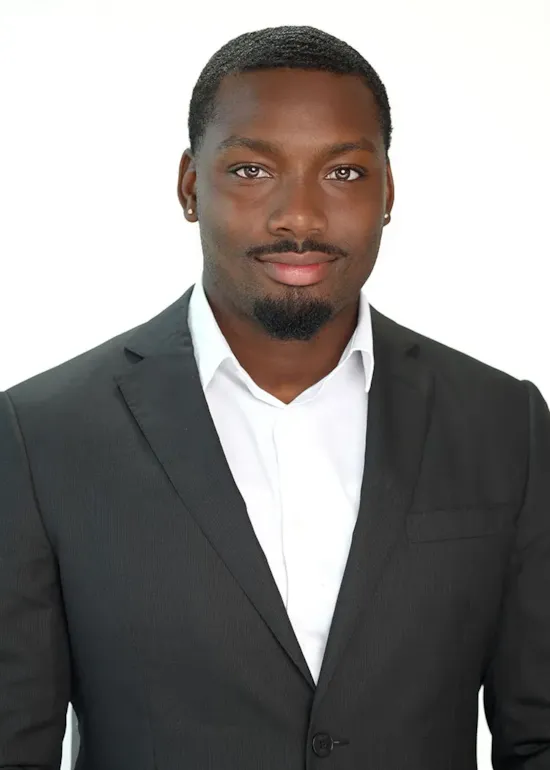 A man in a black suit and white shirt smiles at the camera against a plain white background.