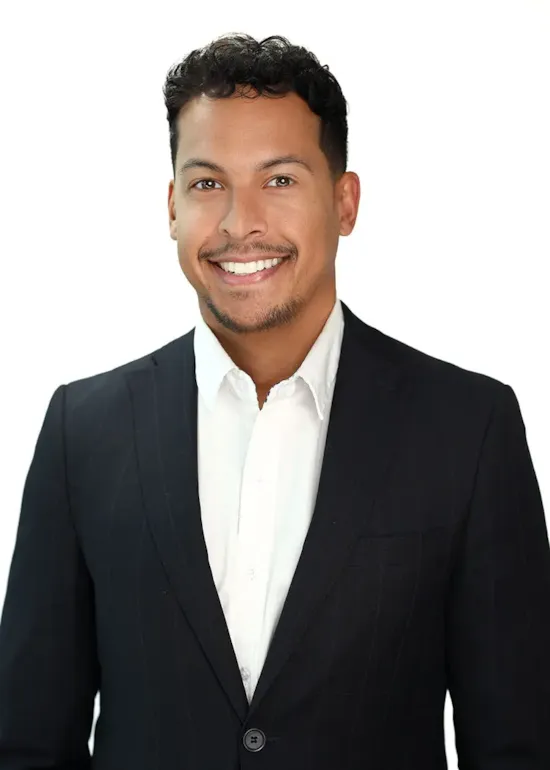 Smiling man with short curly hair in a dark suit jacket and white shirt, standing against a white background.