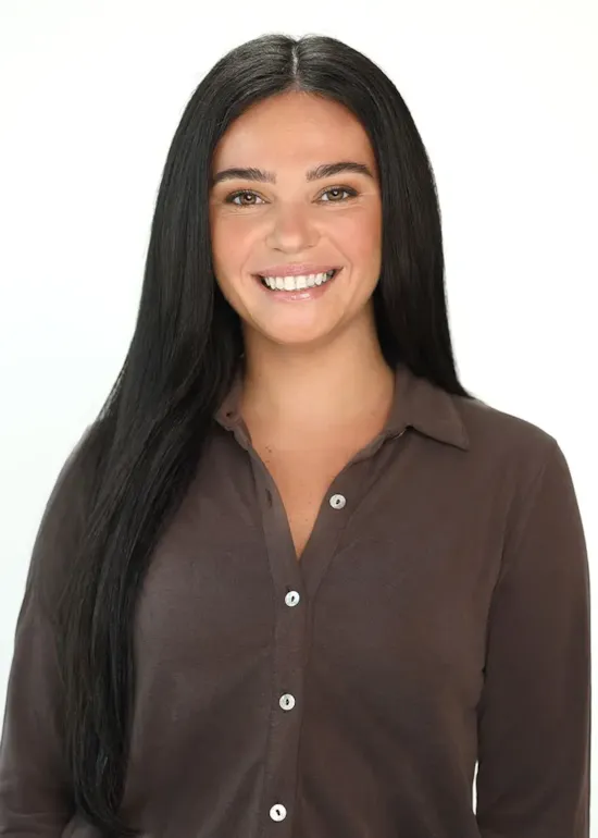 A woman with long dark hair, wearing a brown button-up shirt, smiles at the camera against a plain white background.