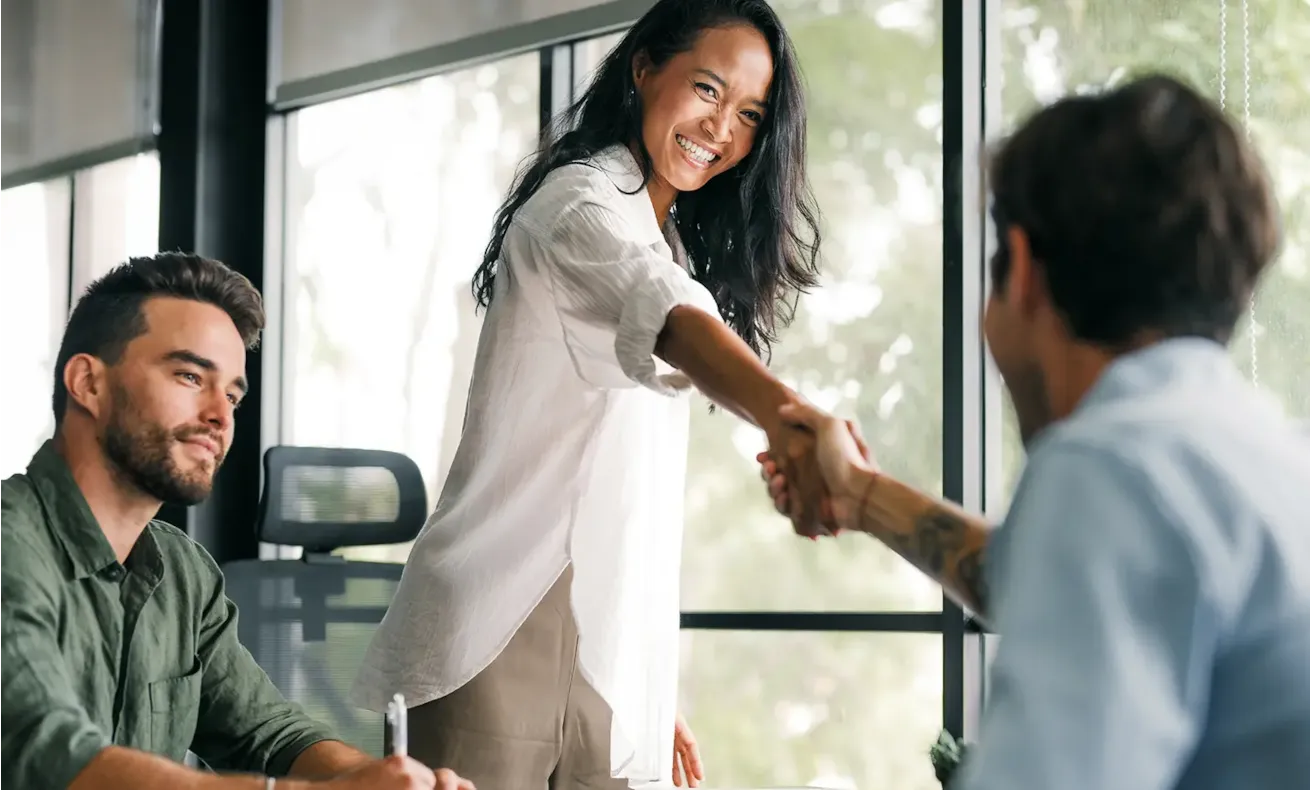 Woman smiling and shaking hands with a seated man in an office, another man watches and smiles.