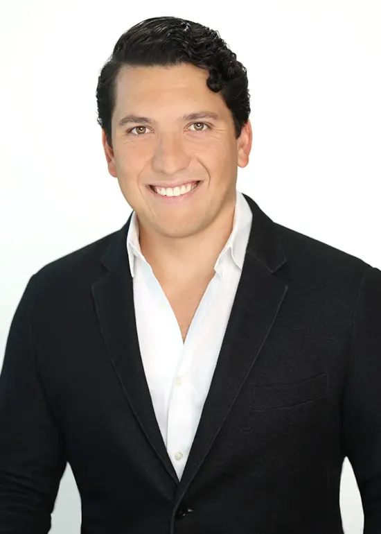 Smiling man with dark hair in a black blazer and white shirt against a plain white background.