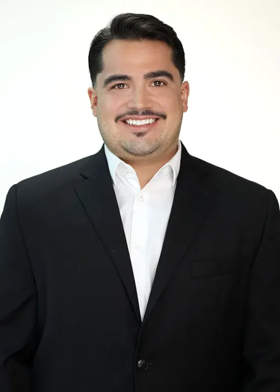 Smiling man in a black suit and white shirt poses against a plain, light background.