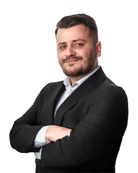 A man in a black suit with folded arms, smiling confidently against a white background.