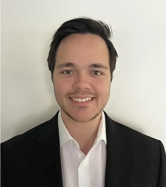 Smiling man with short dark hair wearing a black suit jacket and white shirt, standing against a plain white background.