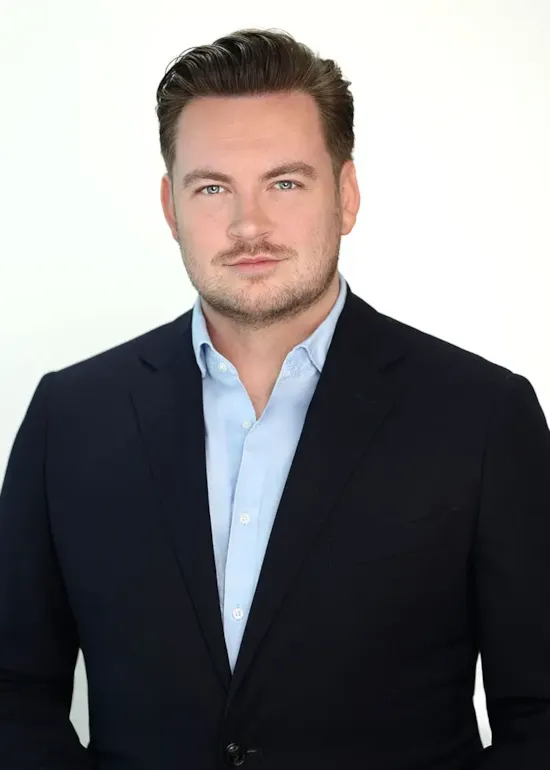 A man in a dark suit and light blue shirt poses against a plain, light background, looking confidently at the camera.