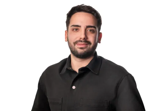 A man with dark hair and beard wearing a black button-up shirt poses against a plain white background.