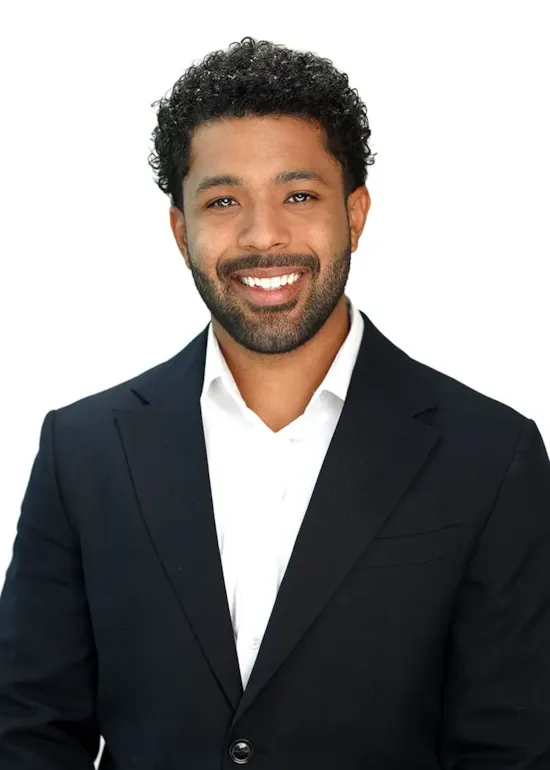 A man with curly hair and a beard smiles, wearing a black blazer and white shirt, against a white background.