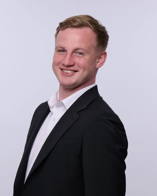 A man in a black suit and white shirt smiles while posing against a plain white background.