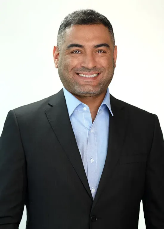 Smiling man in a black suit jacket and blue shirt, standing against a plain light background.