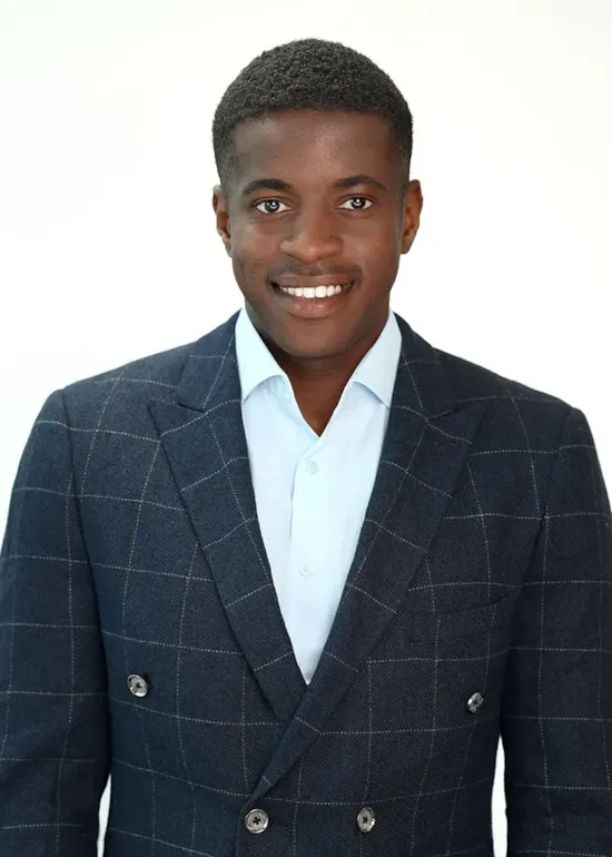 A man in a blue checked suit jacket and light blue shirt smiles at the camera against a white background.