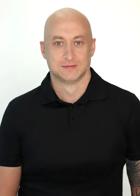 Bald man with light skin in a black polo shirt, looking at the camera against a white background.