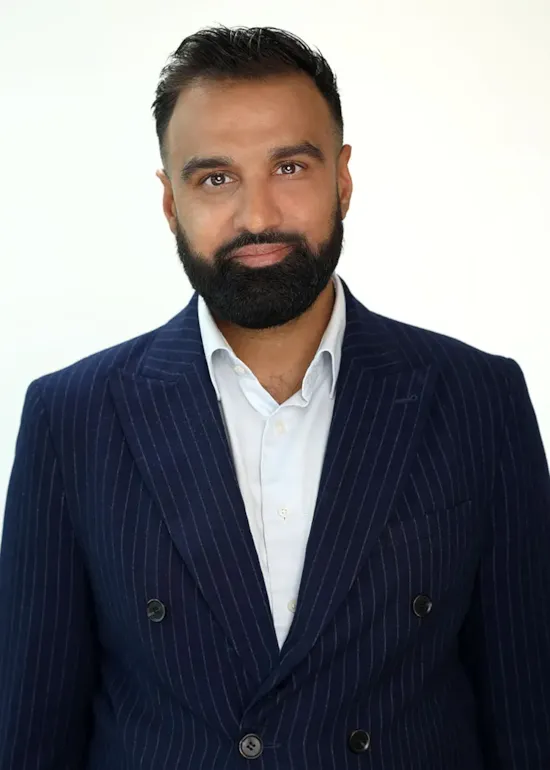 Man with a trimmed beard in a navy pinstripe suit and white shirt, smiling slightly against a plain light background.