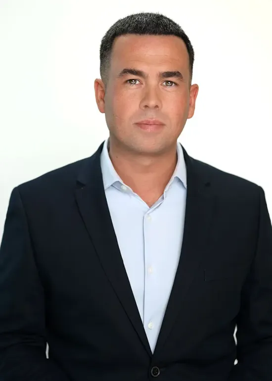 A man in a dark suit jacket and light blue shirt poses against a plain, light background, looking directly at the camera.