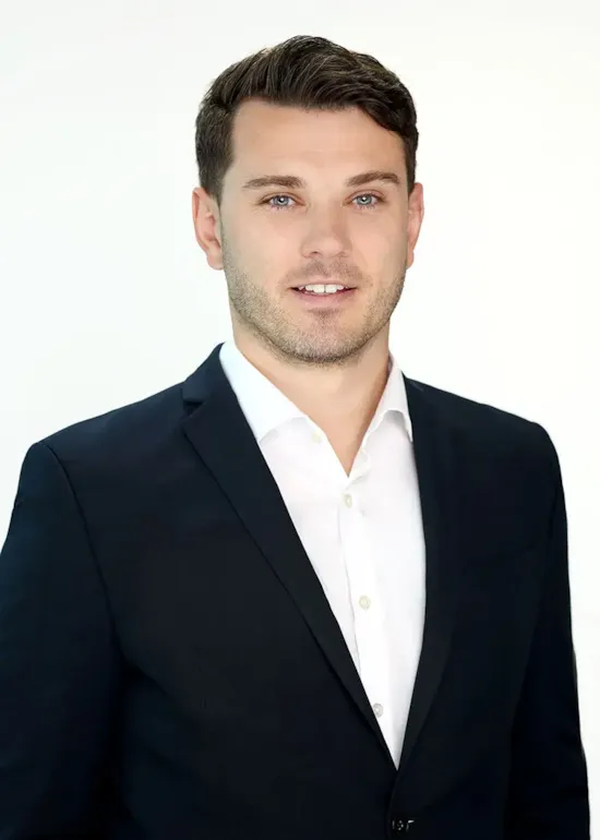 Man in a dark suit and white shirt smiling at the camera against a plain, light background.