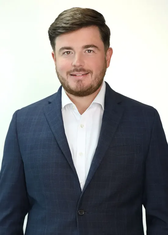A man in a dark checked suit and white shirt smiles in front of a plain light background.