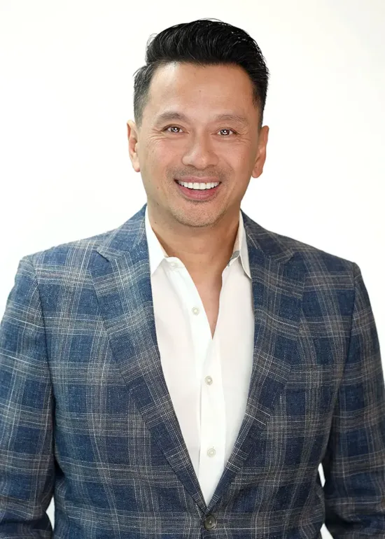 Smiling man with short dark hair wearing a blue plaid blazer and white shirt against a plain light background.