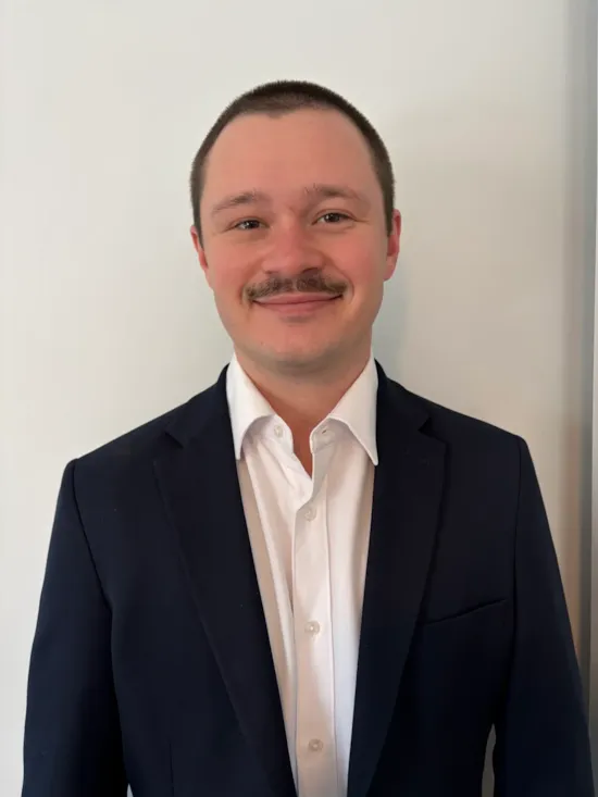 A man in a dark suit and white shirt stands smiling in front of a plain white wall.