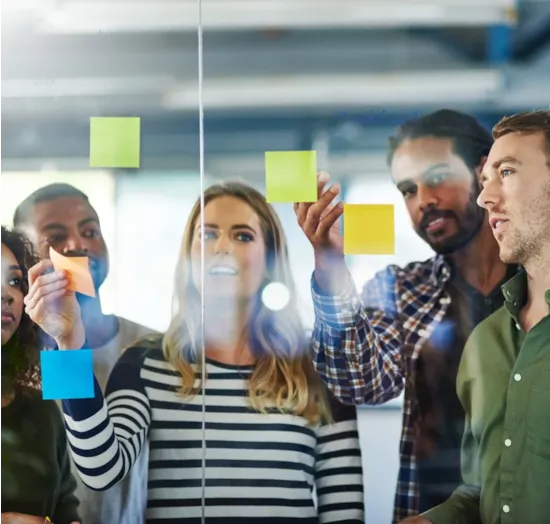 Four people place colorful sticky notes on a glass wall, collaborating and discussing ideas in a modern office setting.