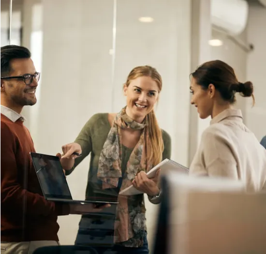 Three people smiling and talking in an office, one holding a laptop and another holding a notebook.