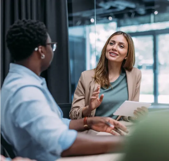 A woman holding a tablet speaks to a colleague during a meeting in a modern office setting.