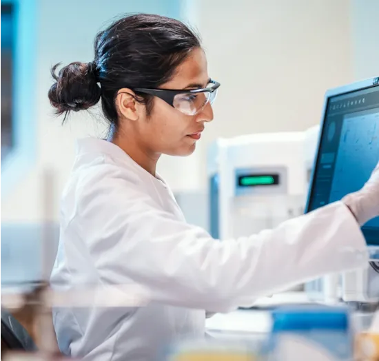 A scientist in a lab coat and safety glasses works on a computer screen in a laboratory setting.