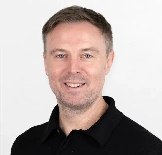 Smiling man with short brown hair and light stubble wearing a black collared shirt against a white background.
