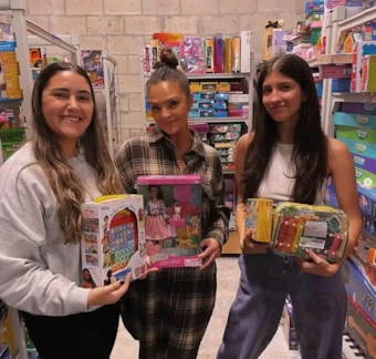 Three women stand in a toy store aisle, smiling and holding various colorful boxed toys.