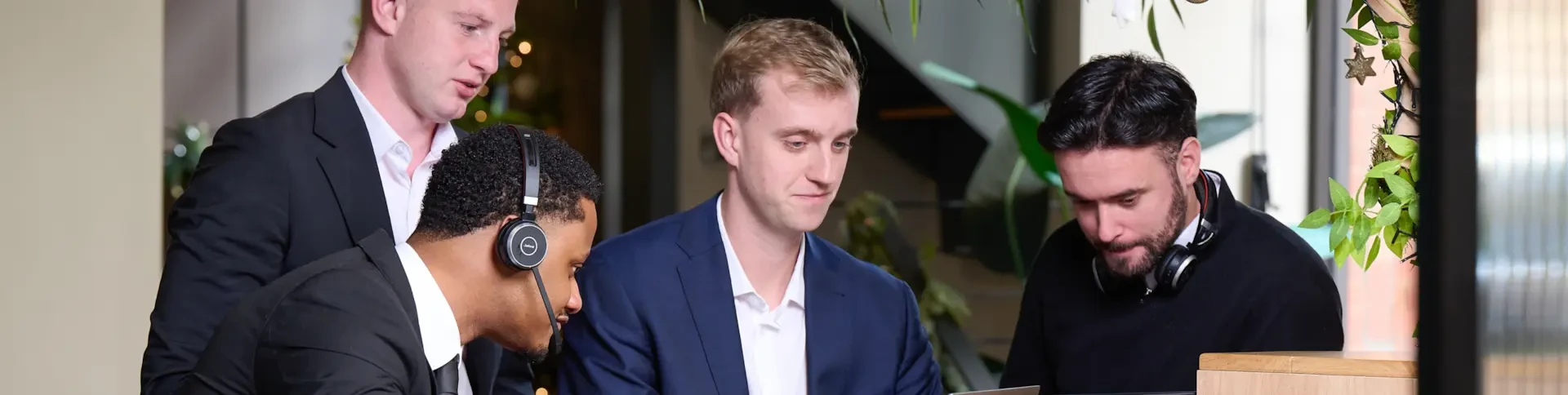 Four men in business attire work together at a desk; two wear headphones and all appear focused on laptops.