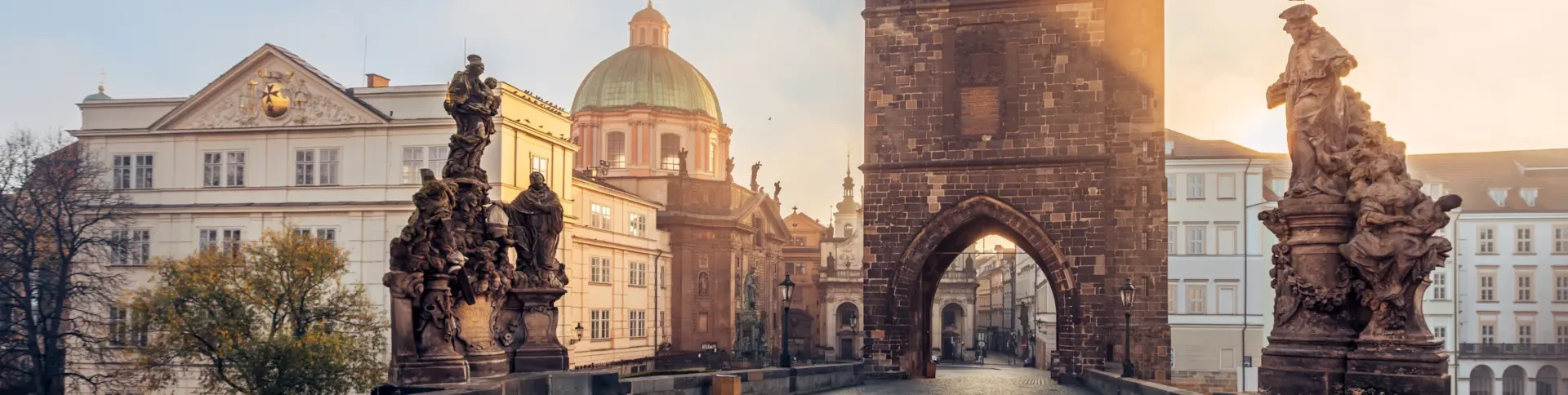 Charles Bridge in Prague at sunrise, featuring statues, historic buildings, and a domed church in soft morning light.