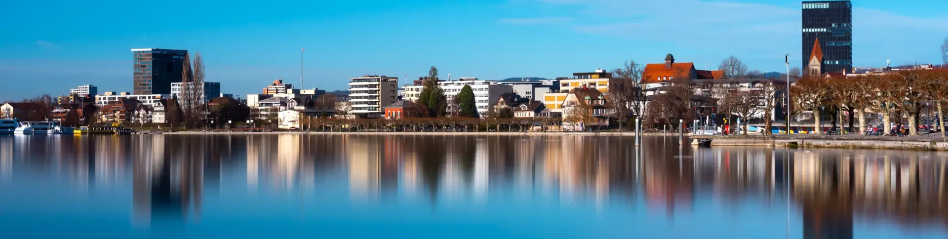City skyline with modern and historic buildings reflected in calm water under a clear blue sky.