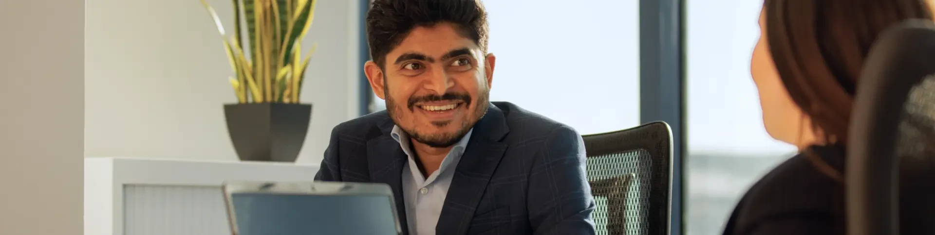 Smiling man in a suit sitting at a desk, talking to a woman in an office with a laptop.
