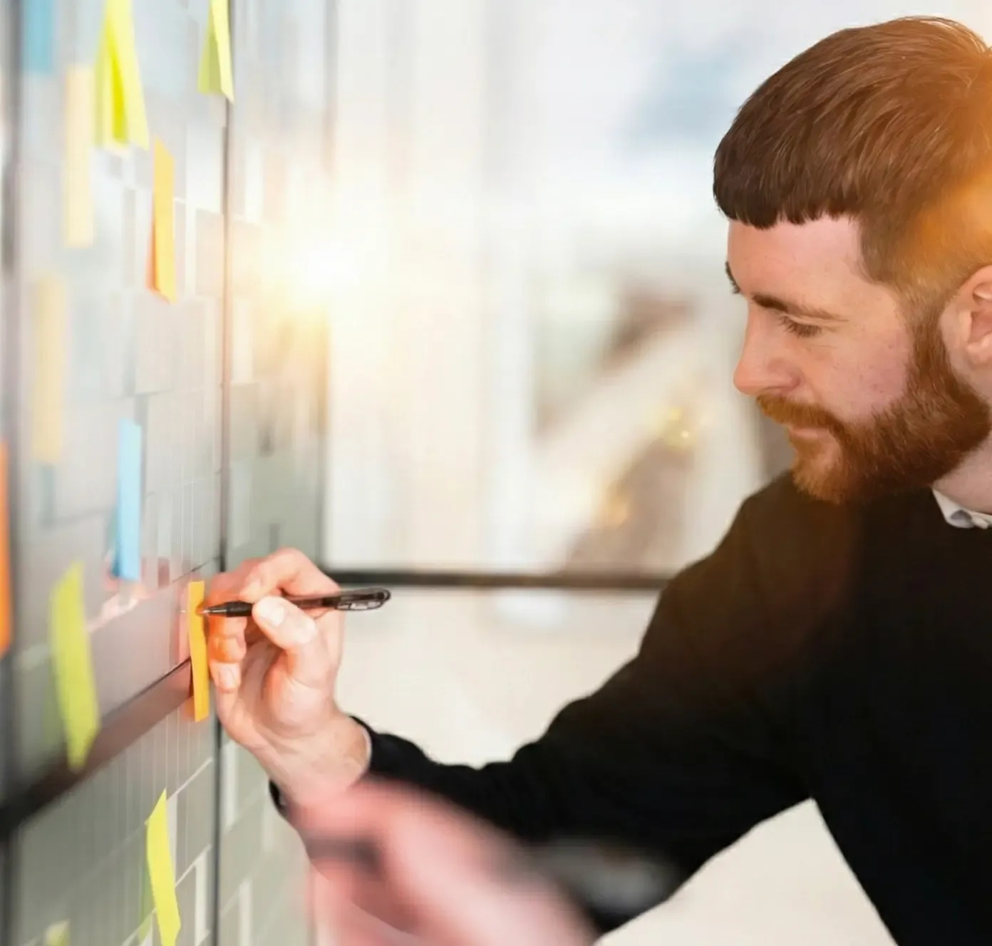 Man writing on colorful sticky notes attached to a glass wall, brainstorming or planning in an office setting.