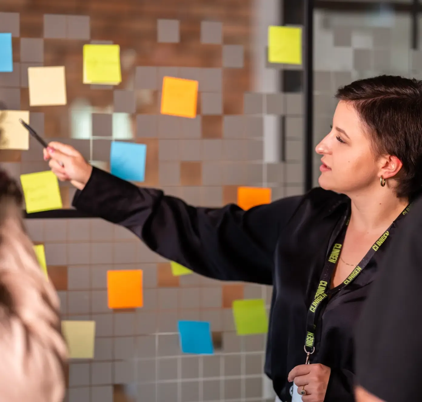 A woman in black points at colorful sticky notes on a glass wall during a meeting or brainstorming session.