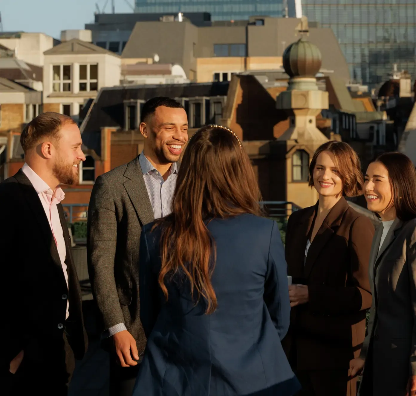 Five people in business attire are smiling and talking on a rooftop with city buildings in the background.