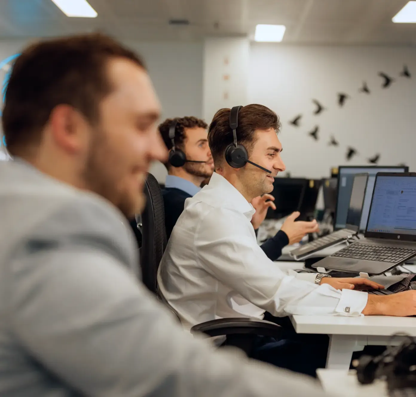 Three men wearing headsets work at computers in a modern office, focused on their tasks and talking to customers.