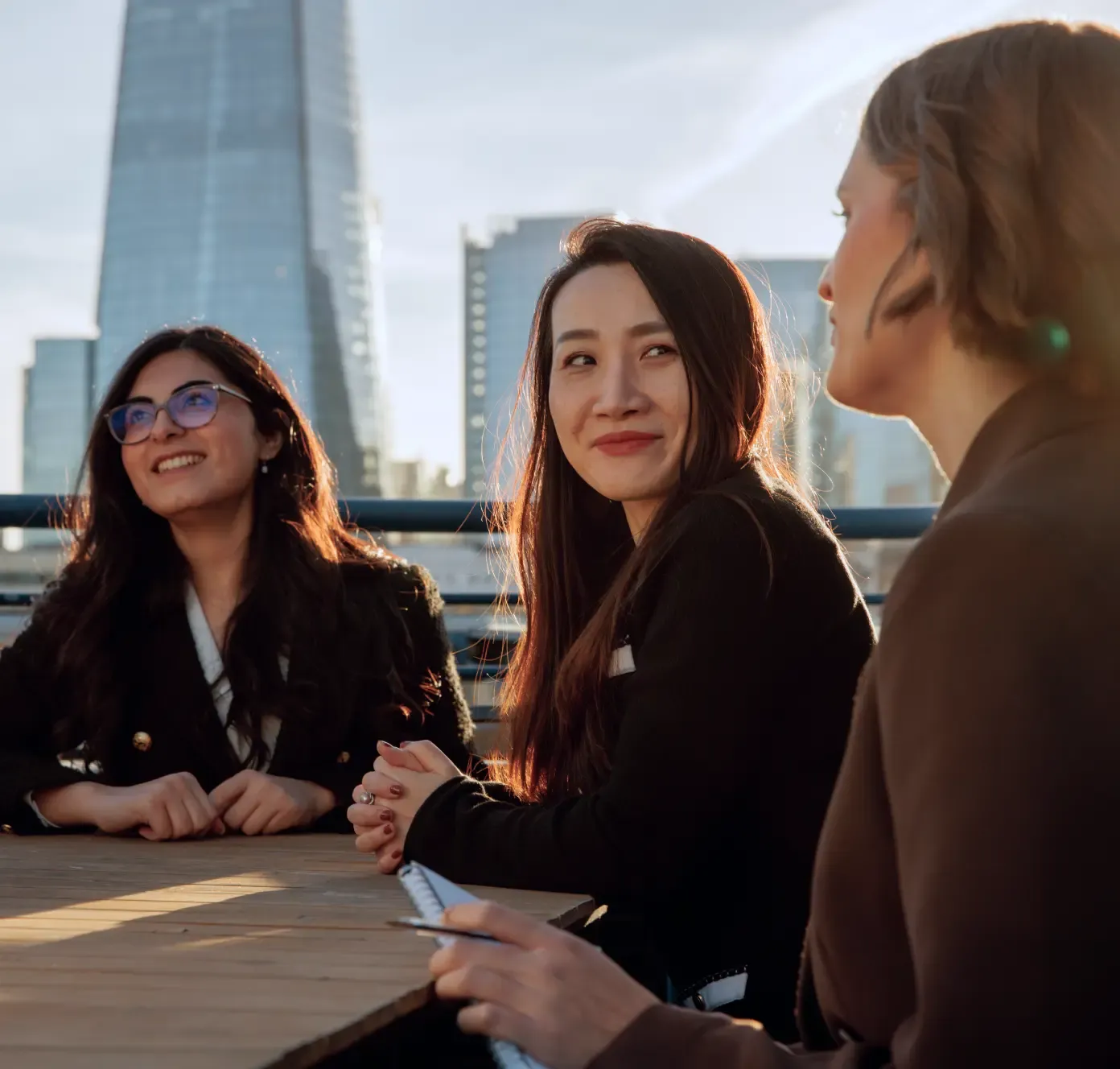 Three women in business attire talk and smile at an outdoor table with city skyscrapers in the background.
