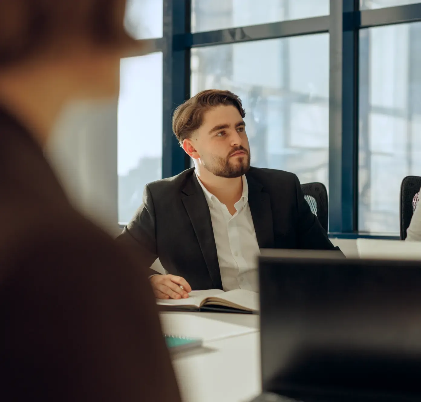 A man in a suit sits at a conference table with a notebook, listening during a meeting in a modern office.