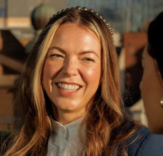 A woman with long hair smiles brightly outdoors, wearing a headband and a white blouse under a blue jacket.