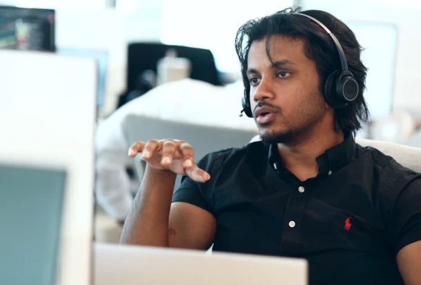 Man wearing a headset gestures with his hand while talking, seated at a desk with computer monitors.