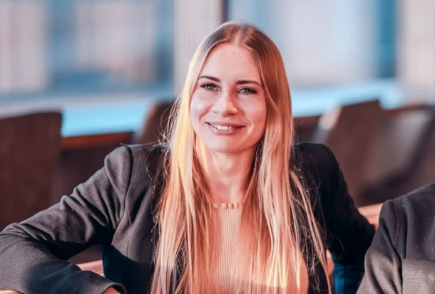 A woman with long blonde hair and a black blazer smiles while sitting indoors in a sunlit room.