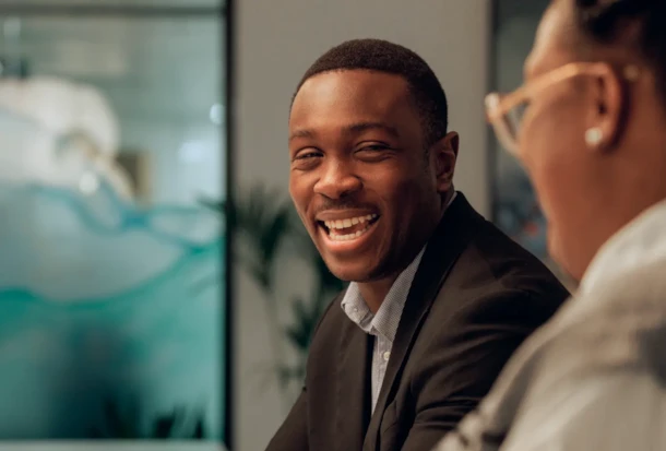 Smiling man in a suit sits at a table, conversing with another person in an office setting.