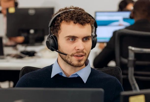 A man with a headset sits at a desk in an office, looking slightly to the side, with computers in the background.