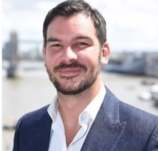 Smiling man in a navy blazer and white shirt standing outdoors with a river and cityscape in the background.