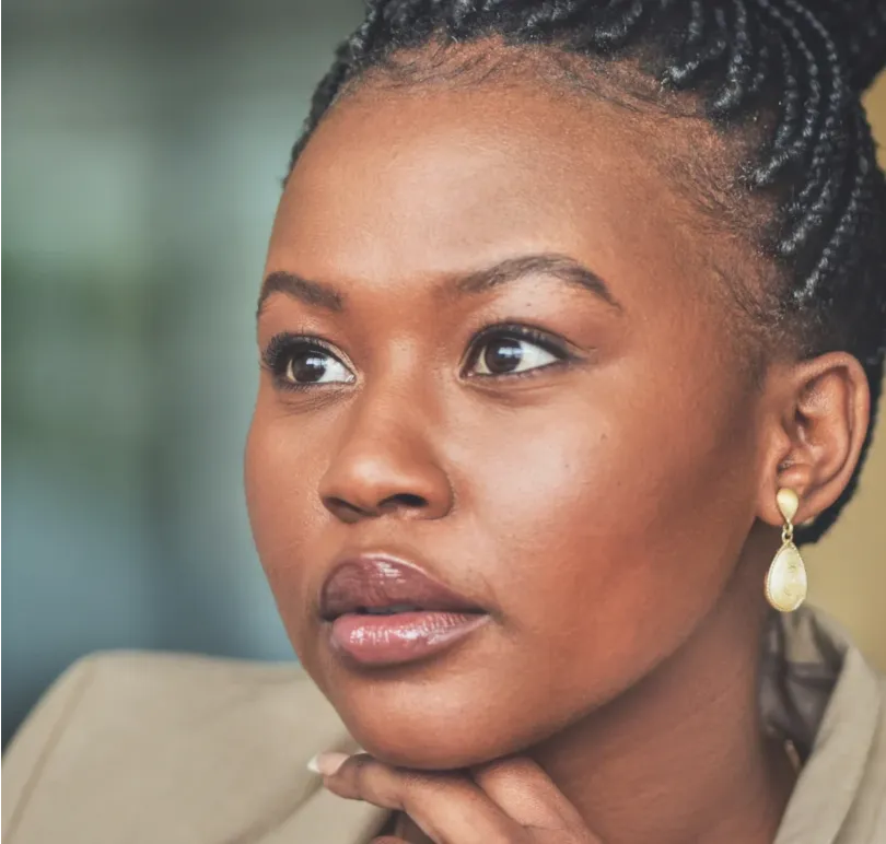 A woman with braided hair and gold earrings looks thoughtfully into the distance, resting her chin on her hand.