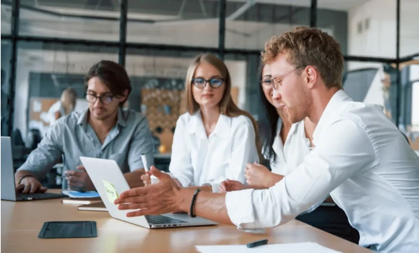 Man gesturing while explaining something on a laptop to two colleagues during a meeting in a modern office.