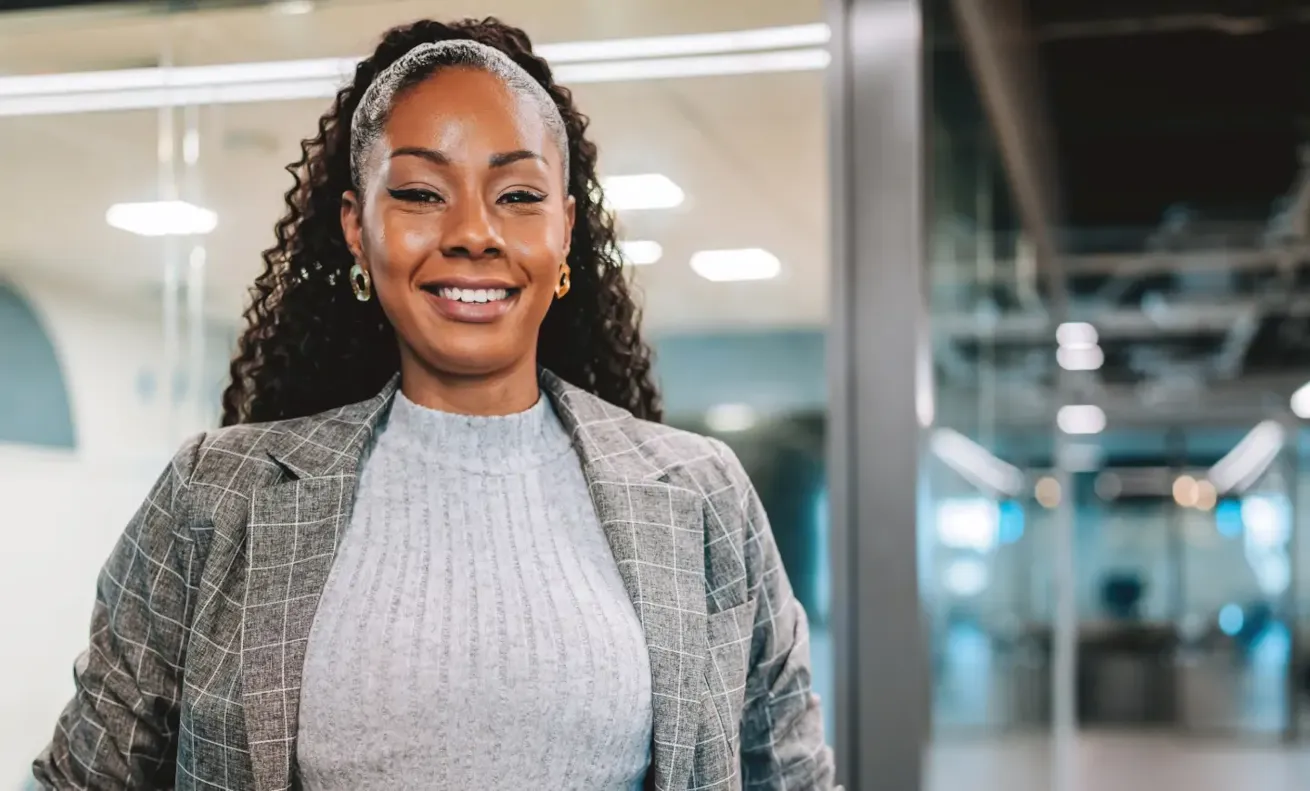 Smiling professional woman in a gray plaid blazer standing confidently in a modern office hallway.